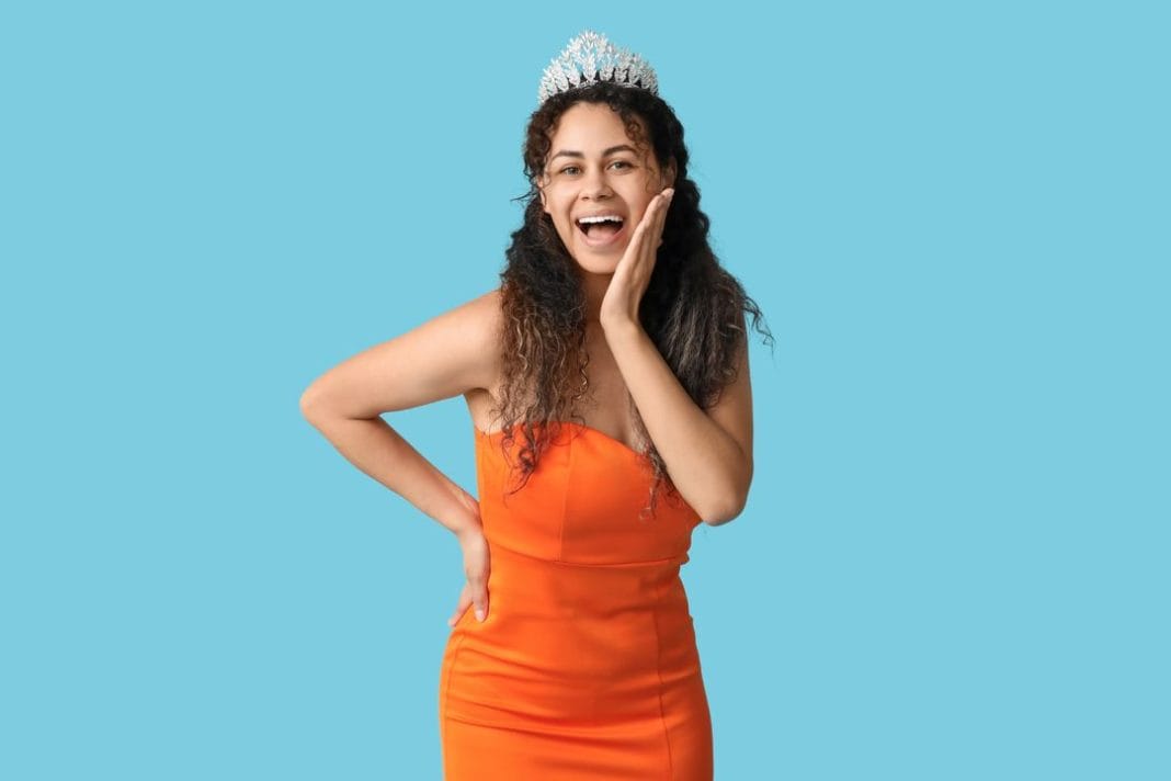 Happy young African-American woman in stylish orange prom dress and tiara on blue background femme en robe de soirée