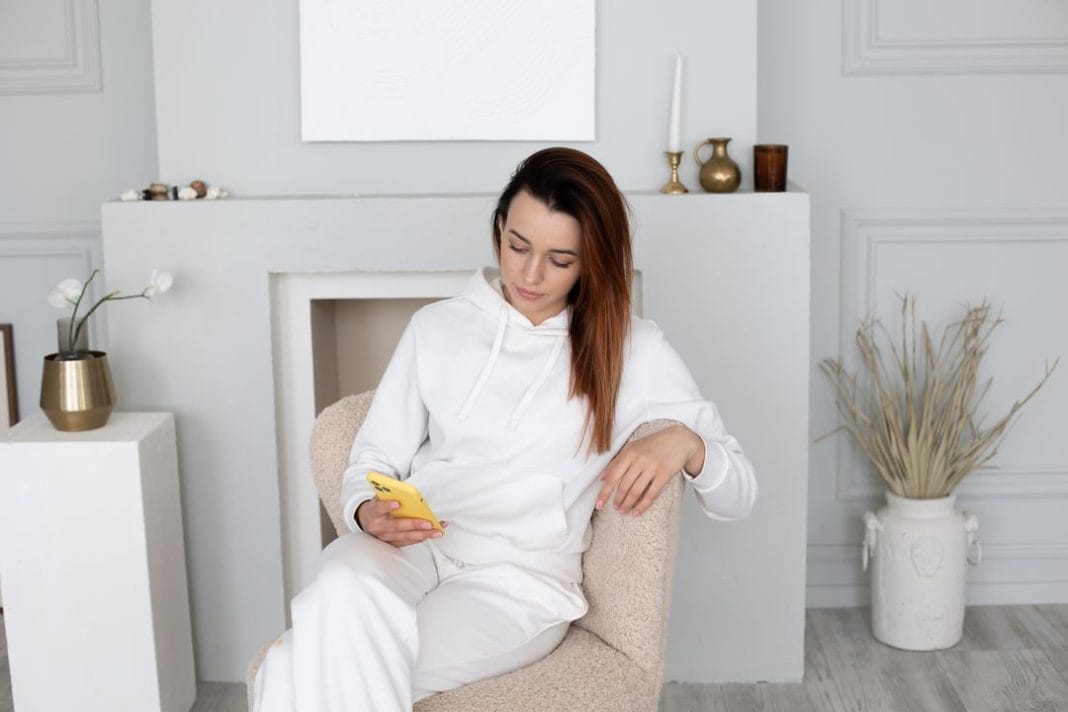 Woman in a white tracksuit sits on an armchair and watches the Internet on her phone.